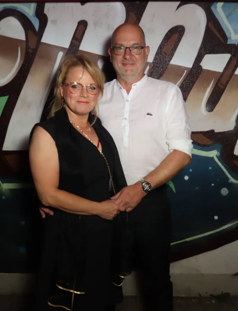 Ein Paar steht nebeneinander und hält sich an den Händen, beide tragen Brille. Im Hintergrund eine Wand mit Graffiti.