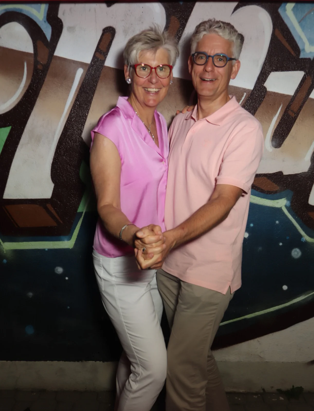 Ein Paar tanzt eng umschlungen, beide tragen Brillen und farblich abgestimmte Kleidung. Im Hintergrund eine Graffiti-Wand.