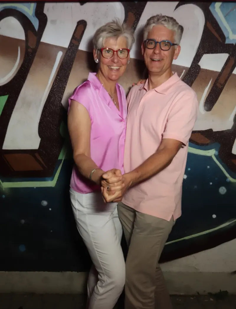 Ein Paar tanzt eng umschlungen, beide tragen Brillen und farblich abgestimmte Kleidung. Im Hintergrund eine Graffiti-Wand.