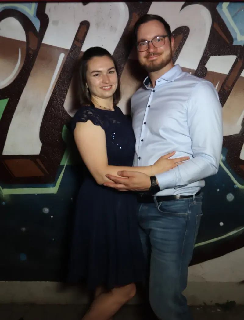 Ein Paar steht eng umschlungen, die Frau trägt ein dunkelblaues Kleid, der Mann ein helles Hemd. Im Hintergrund Graffiti.