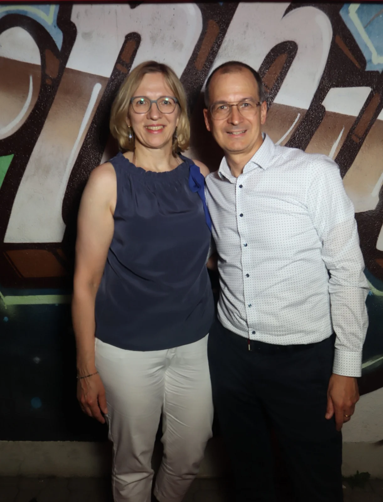 Ein Paar steht nebeneinander, beide tragen Brille und helle Kleidung. Im Hintergrund ist eine bemalte Wand mit Graffiti.