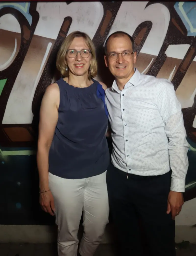 Ein Paar steht nebeneinander, beide tragen Brille und helle Kleidung. Im Hintergrund ist eine bemalte Wand mit Graffiti.