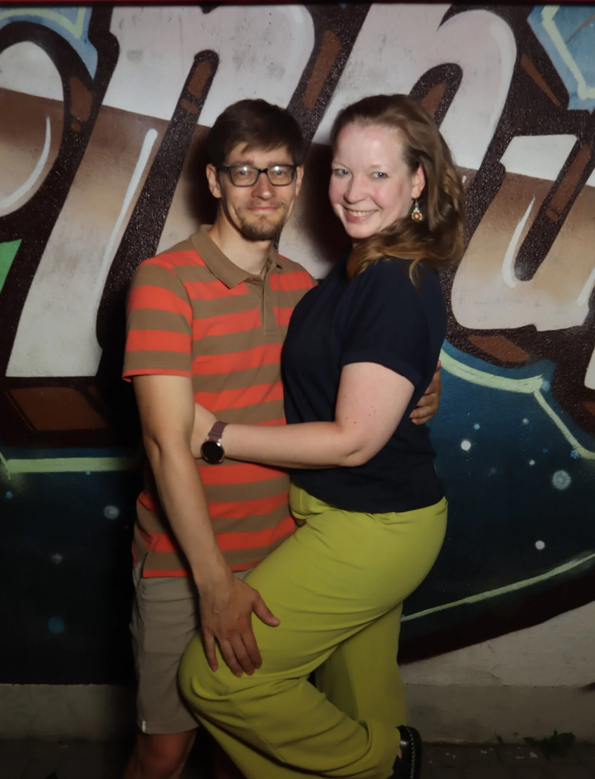 Ein Paar posiert eng umschlungen, die Frau trägt gelbe Hose und hebt ein Bein. Im Hintergrund eine bemalte Wand mit Graffiti.