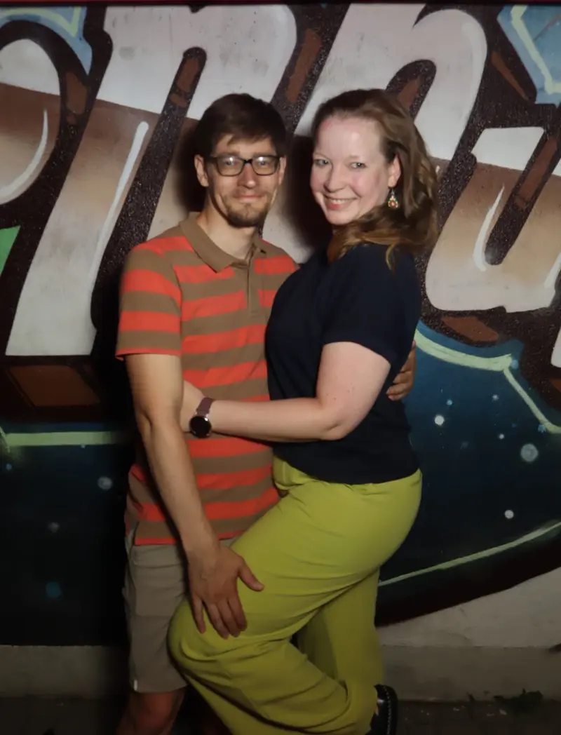 Ein Paar posiert eng umschlungen, die Frau trägt gelbe Hose und hebt ein Bein. Im Hintergrund eine bemalte Wand mit Graffiti.