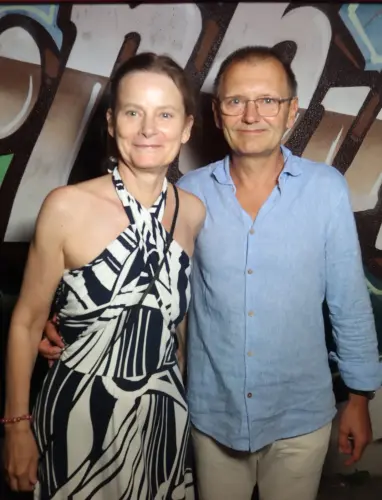Ein Paar steht nebeneinander, die Frau trägt ein gemustertes Kleid, der Mann ein hellblaues Hemd. Im Hintergrund Graffiti.