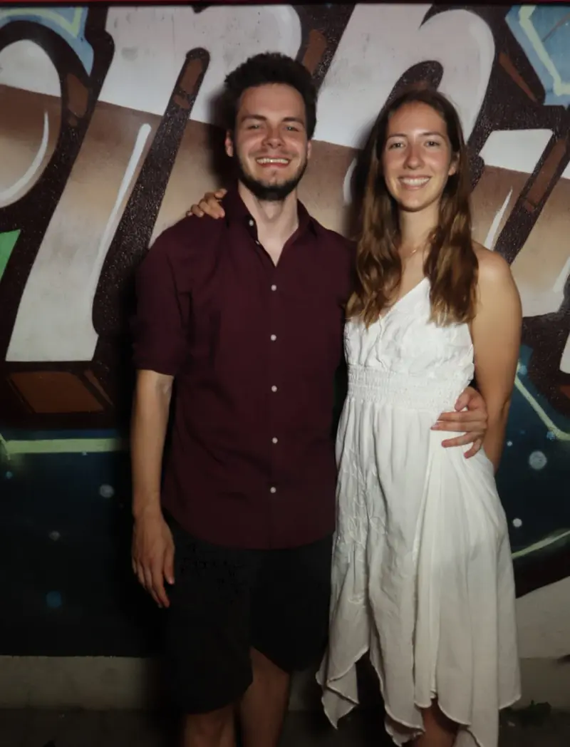 Ein junges Paar steht nebeneinander, der Mann trägt ein Hemd, die Frau ein weißes Kleid. Im Hintergrund Graffiti.