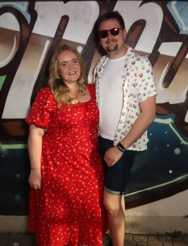 Ein Mann und eine Frau posieren vor einer bemalten Wand. Beide tragen sommerliche Kleidung mit Muster.