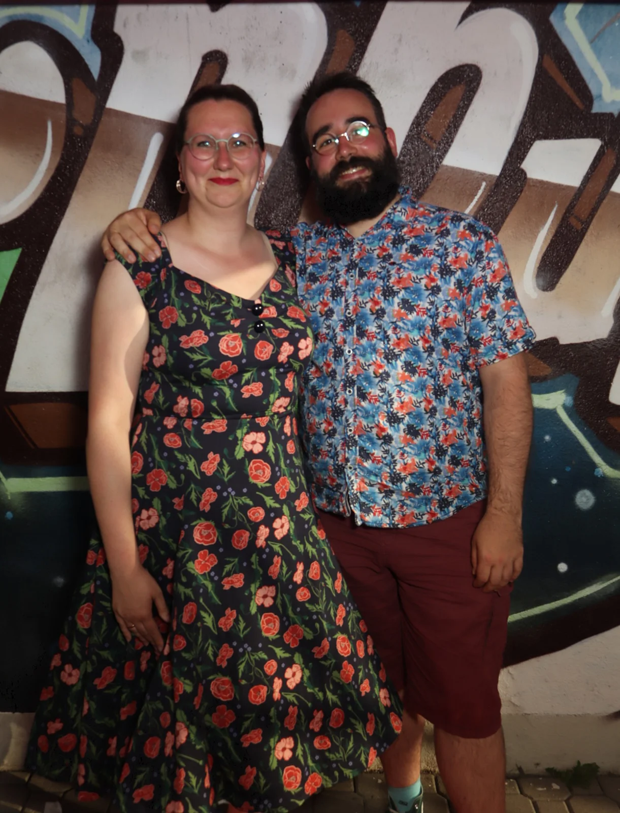 Zwei Personen stehen nebeneinander und blicken in die Kamera. Im Hintergrund eine bemalte Wand.