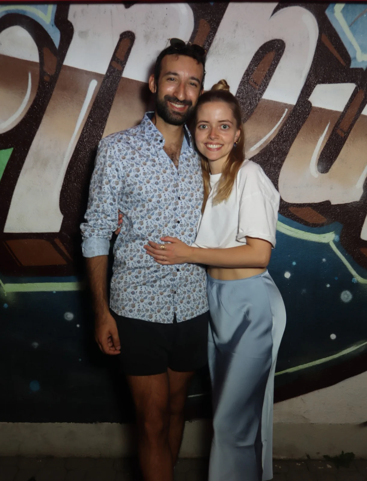 Ein Paar steht nebeneinander, der Mann trägt ein gemustertes Hemd, die Frau ein weißes Shirt. Im Hintergrund Graffiti.