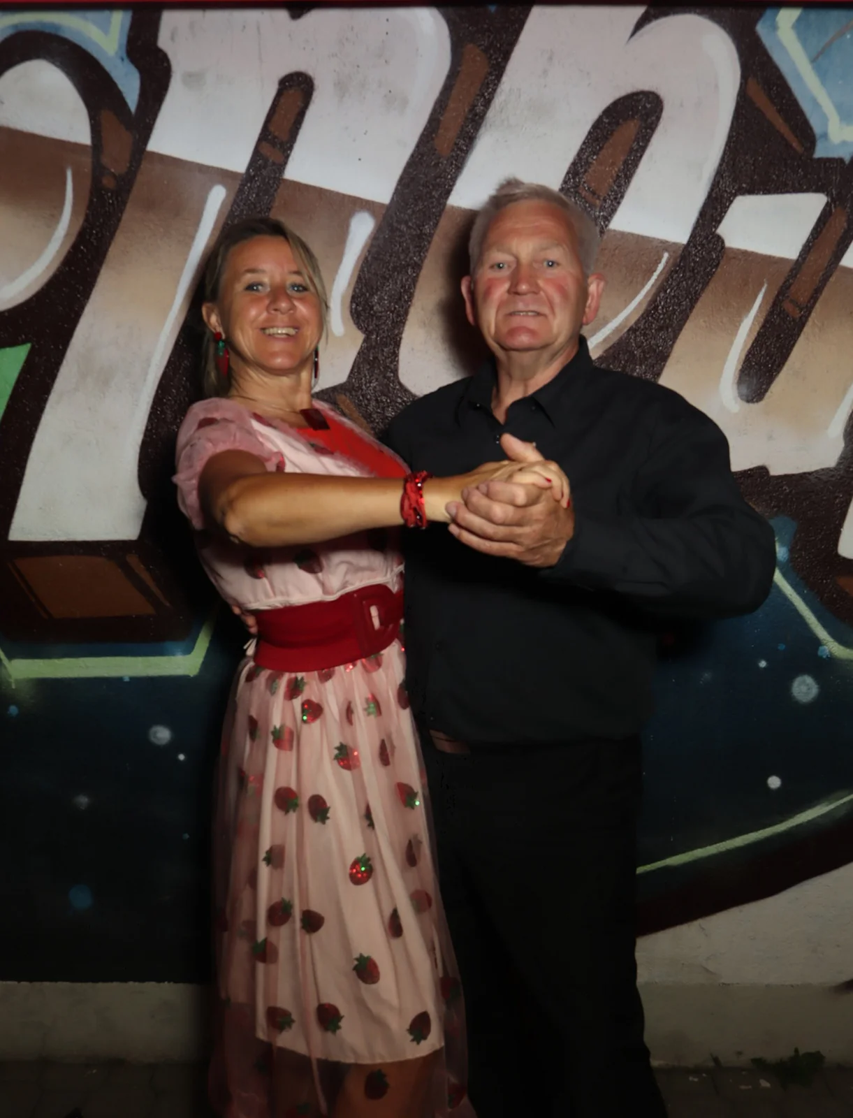 Ein Paar steht in Tanzhaltung, die Frau trägt ein Kleid mit Erdbeermuster, der Mann ein Hemd. Im Hintergrund Graffiti.