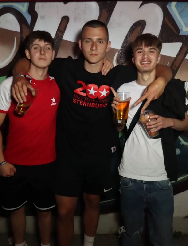 Drei junge Männer stehen nebeneinander, jeder hält ein Getränk in der Hand. Im Hintergrund eine Wand mit Graffiti-Motiv.