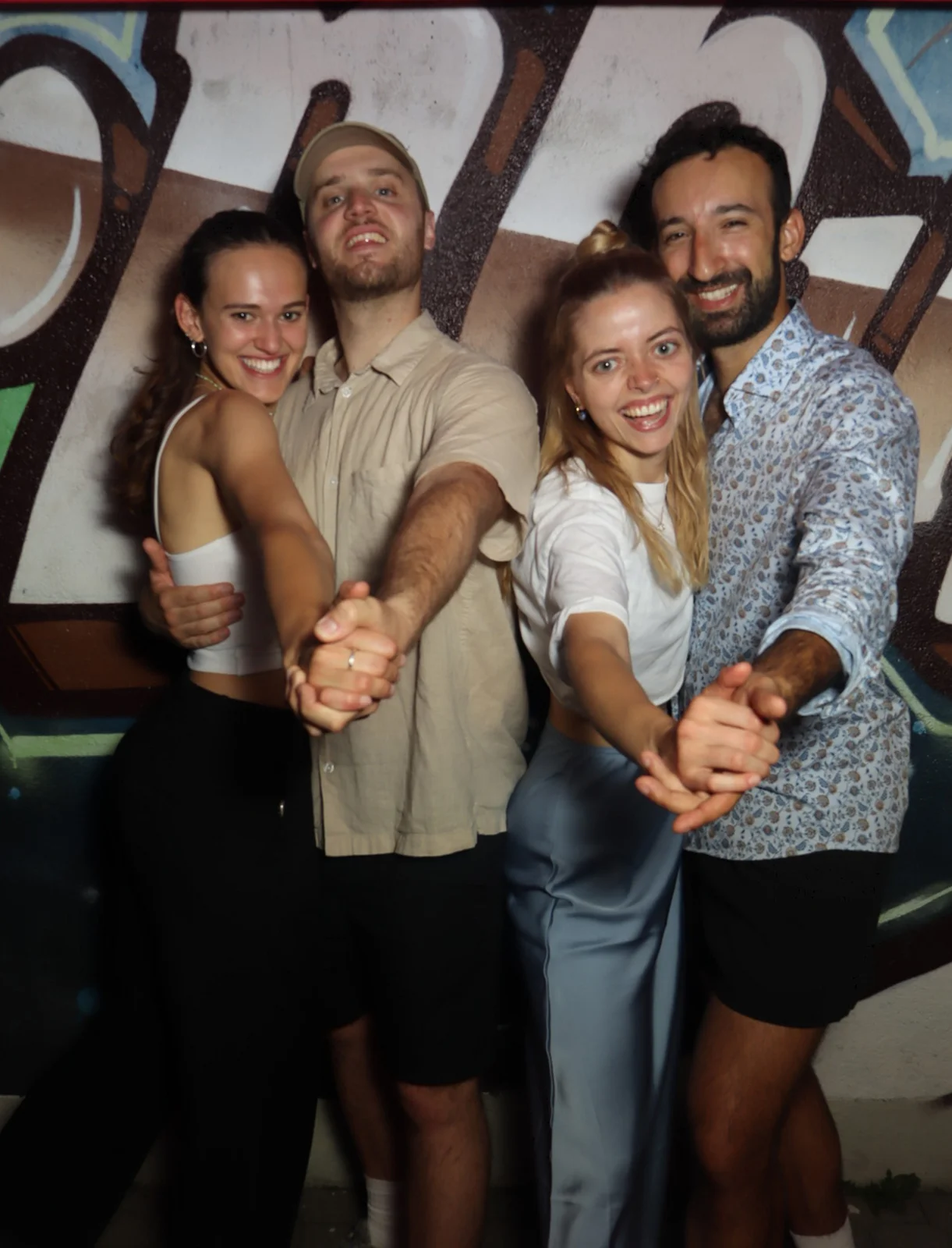 Zwei Paare stehen nebeneinander und halten sich tanzend an den Händen. Im Hintergrund eine Wand mit Graffiti-Schriftzug.