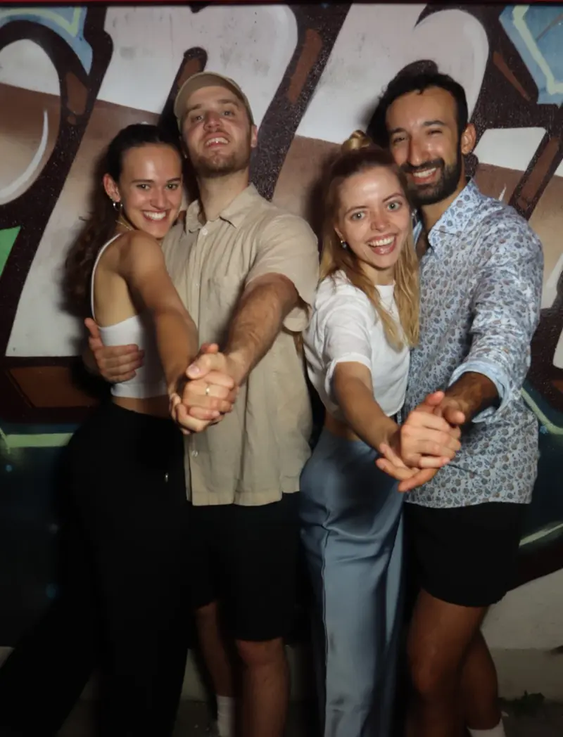 Zwei Paare stehen nebeneinander und halten sich tanzend an den Händen. Im Hintergrund eine Wand mit Graffiti-Schriftzug.