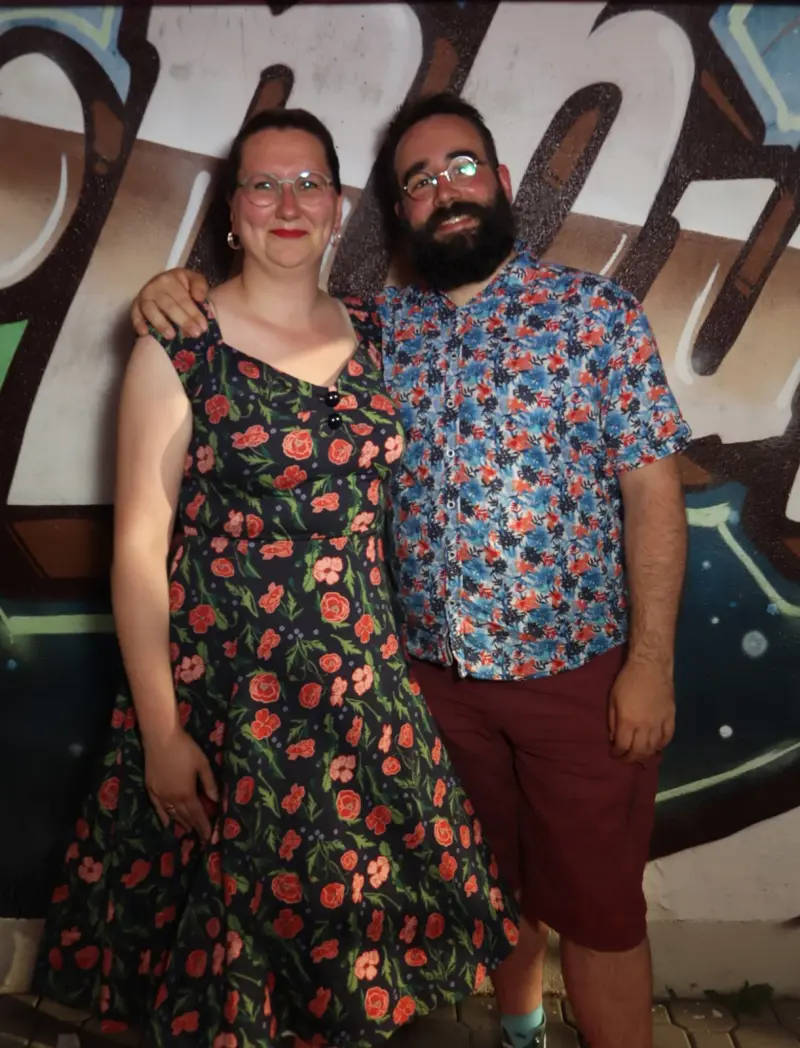 Zwei Personen stehen nebeneinander und blicken in die Kamera. Im Hintergrund eine bemalte Wand.