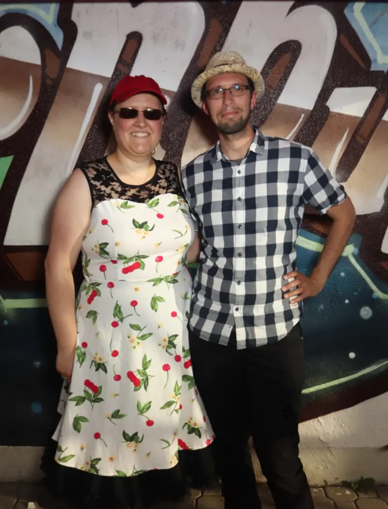 Ein Mann und eine Frau stehen nebeneinander und schauen in die Kamera. Im Hintergrund eine bunt bemalte Wand.