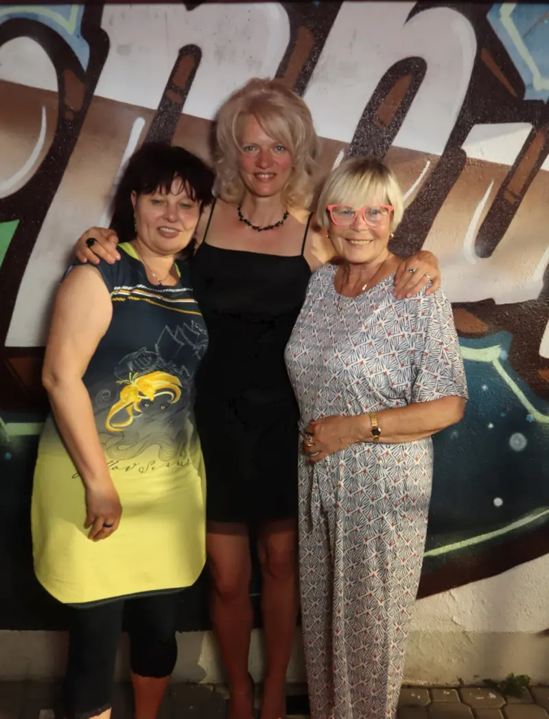 Drei Frauen stehen nebeneinander und lächeln in die Kamera. Im Hintergrund eine bunt bemalte Wand mit Buchstaben.