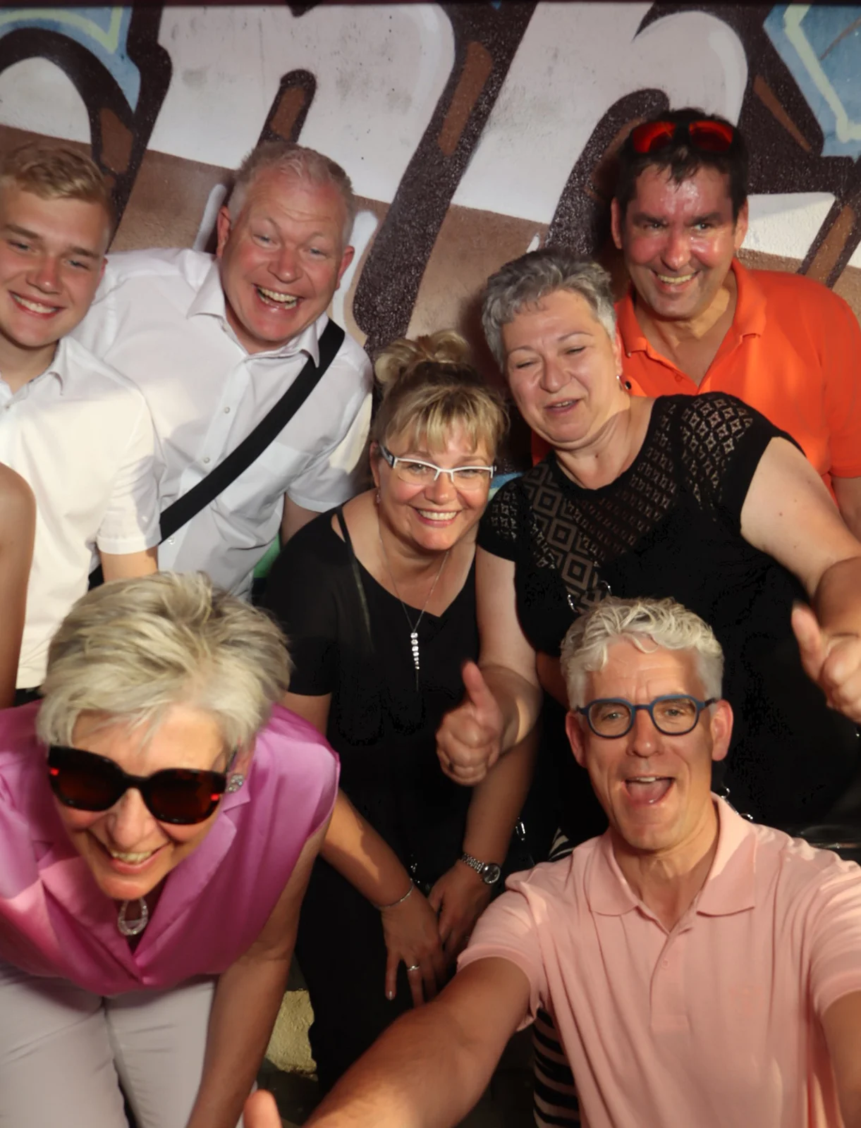 Acht Personen posieren lachend dicht beieinander für ein Gruppenfoto. Im Hintergrund eine Wand mit Graffiti-Schriftzug.