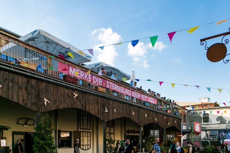 Menschen feiern auf Terrasse mit Sternburg-Banner. Bunte Wimpel schaffen sommerliche Festivalstimmung.