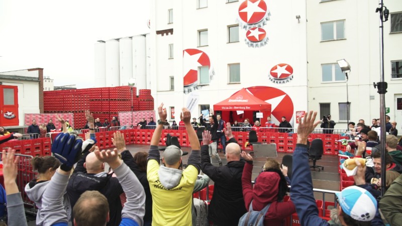 Mehrere Menschen jubeln bei einem Open-Air-Event vor einem Zelt und roten Stapelboxen.