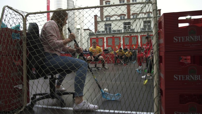 Eine Person sitzt auf einem Bürostuhl hinter einem Netz und spielt Hockey mit einer Gruppe junger Menschen.