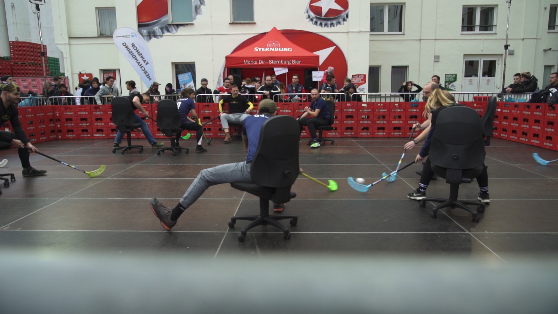 Mehrere Personen spielen in einem umzäunten Bereich Hockey auf Bürostühlen.