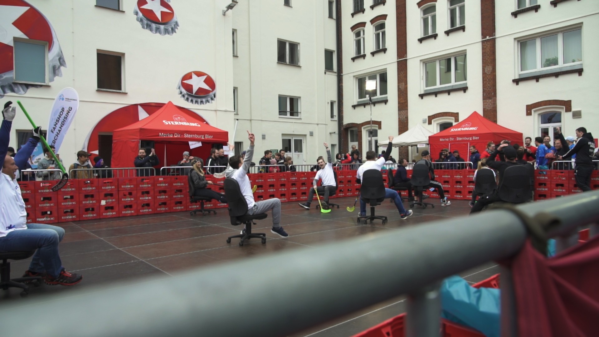 Menschen nehmen an einem Bürostuhlrennen auf einem Platz teil, umgeben von Zuschauern.