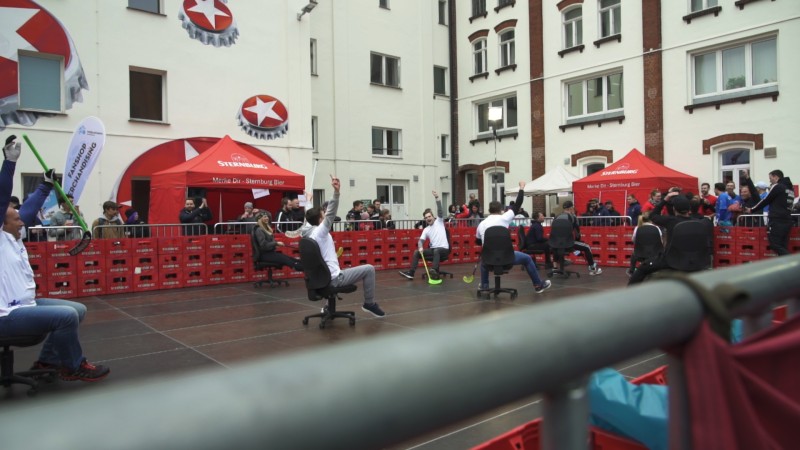 Menschen nehmen an einem Bürostuhlrennen auf einem Platz teil, umgeben von Zuschauern.