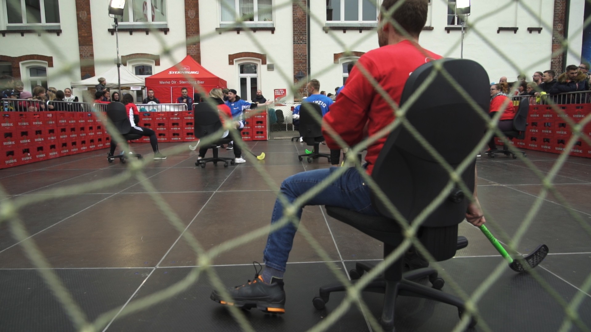 Menschen spielen in einem Innenhof Bürostuhl-Hockey, umgeben von Zuschauenden.