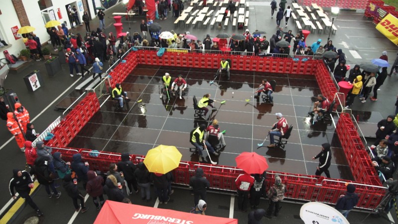Menschen bei einem Stuhlwettrennen auf einem nassen Platz, umgeben von Zuschauern mit Schirmen.