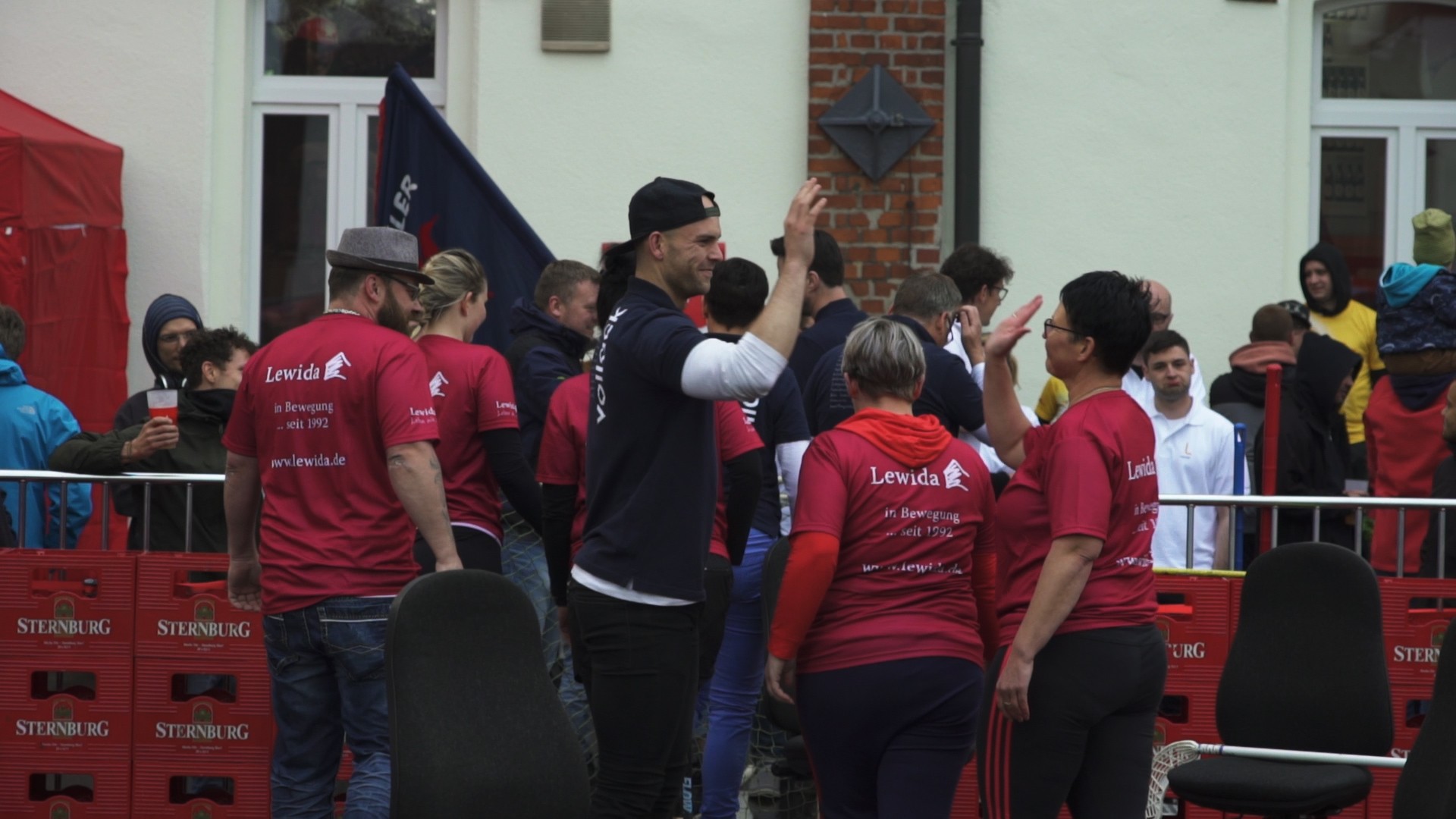 Mehrere Personen in roten Oberteilen stehen draußen bei einer Veranstaltung. Ein Mann winkt lächelnd.