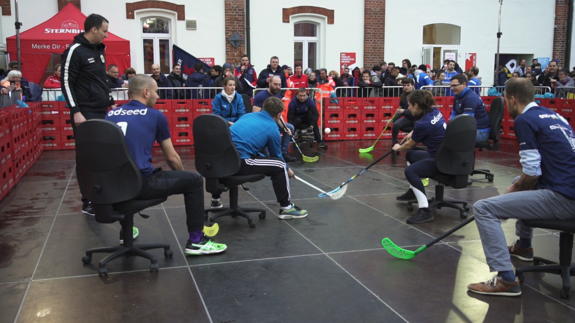 Vier Personen spielen in Bürostühlen auf einer Fläche Hockey, umgeben von Zuschauern.