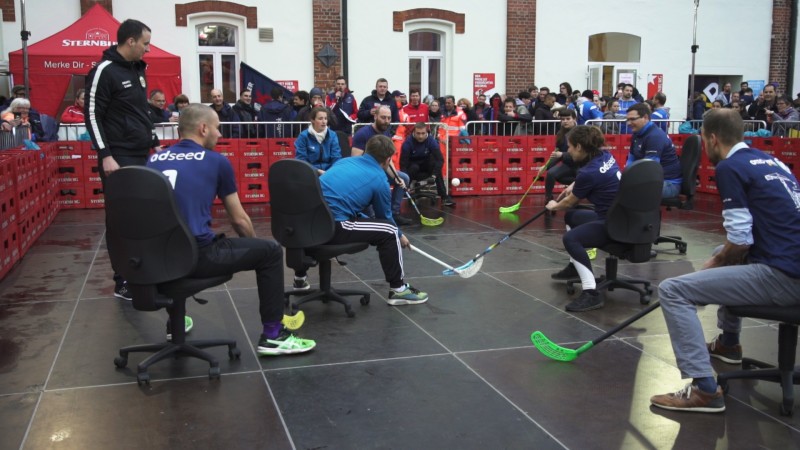 Vier Personen spielen in Bürostühlen auf einer Fläche Hockey, umgeben von Zuschauern.