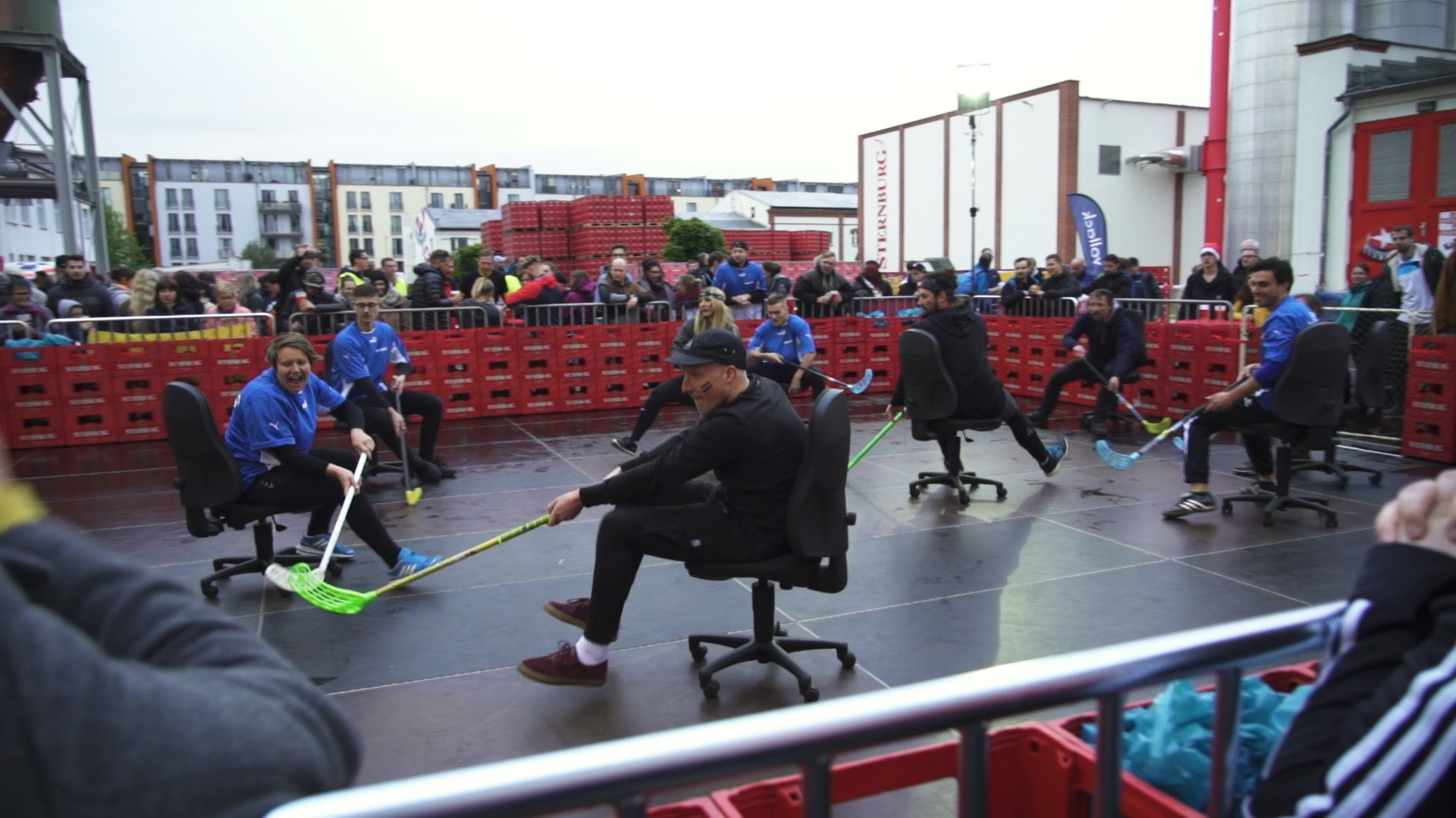Menschen spielen in Bürostühlen Hockey auf einem Platz voller Zuschauer.