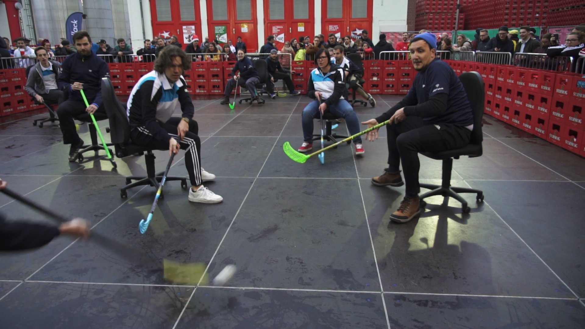 Mehrere Personen spielen in einer Halle mit Stühlen Hockey. Sie verwenden bunte Hockeyschläger, um einen Ball zu spielen.