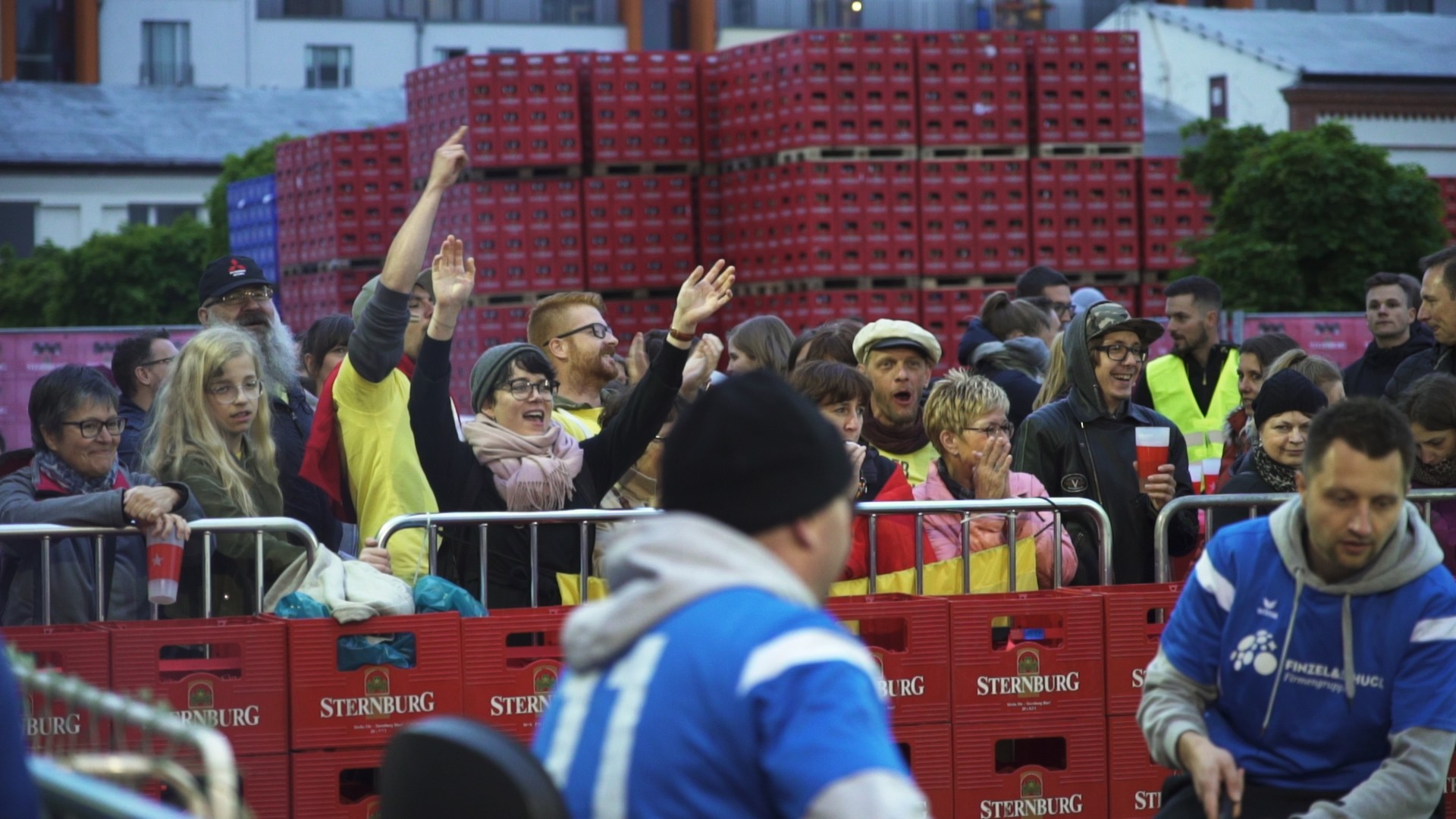 Eine Menschenmenge steht hinter Absperrgittern, einige winken und jubeln.