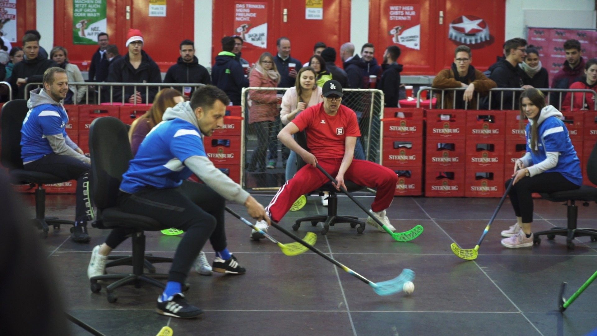 Vier Personen spielen auf Bürostühlen in einer Halle ein Ballspiel mit Schlägern.