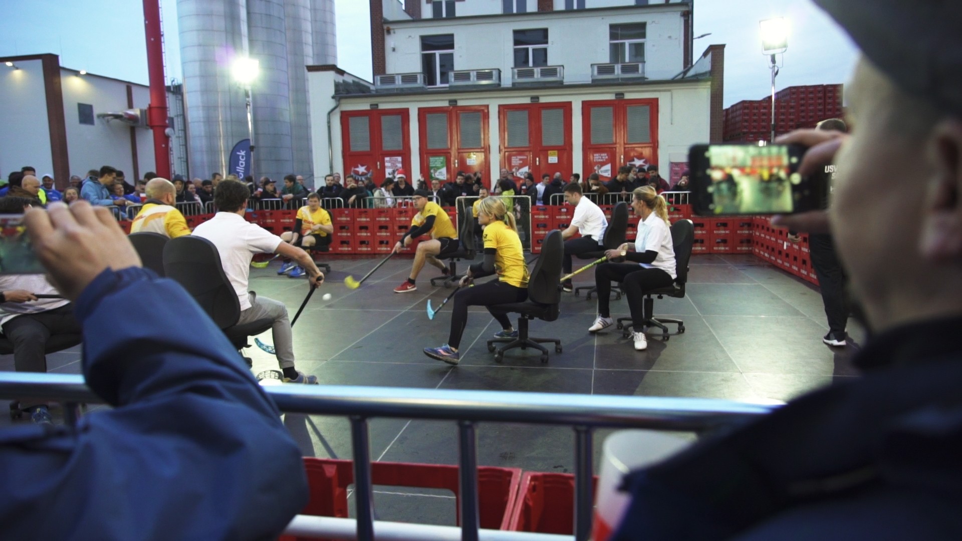 Menschen spielen auf Bürostühlen sitzend Hockey vor einem Publikum im Freien.