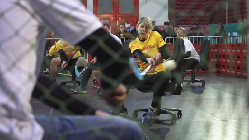 Eine Frau im gelben Shirt fährt auf einem Bürostuhl und nutzt einen Besen als Paddel.