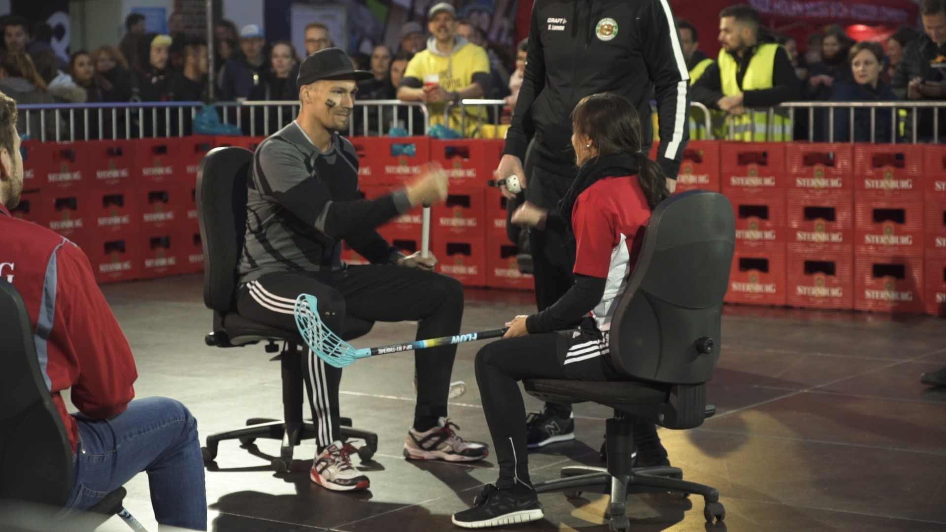 Ein Mann und eine Frau sitzen auf Bürostühlen und spielen mit Schlägern einen Ball. Sie tragen sportliche Kleidung.