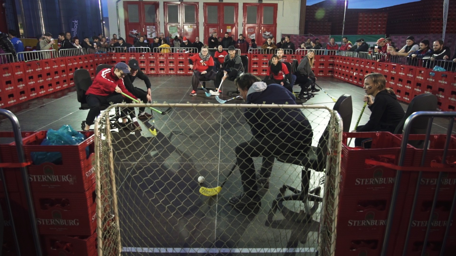 Zwei Teams spielen in einer improvisierten Arena Hockey, Zuschauer beobachten das Spiel.