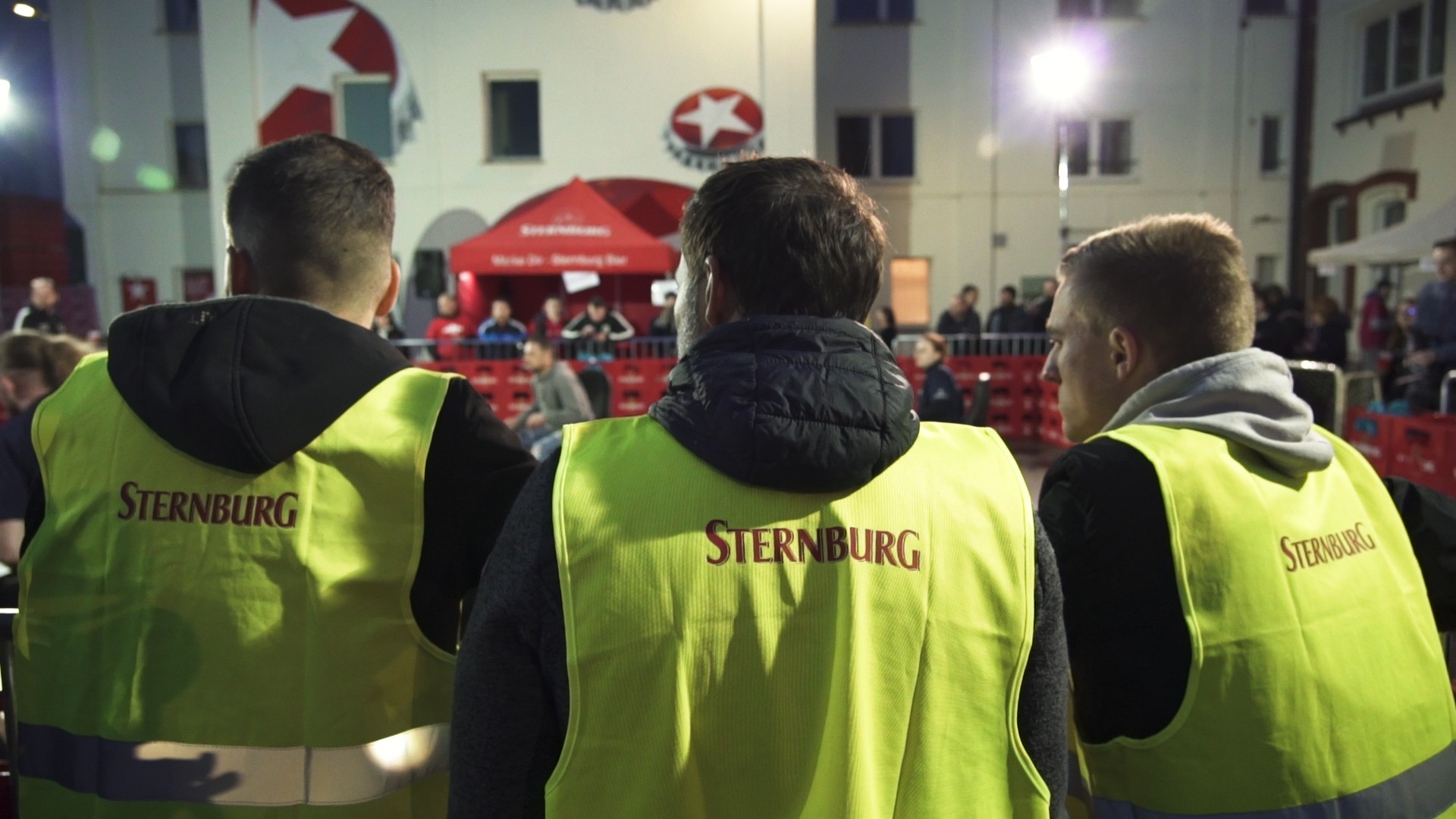 Drei Personen in neongelben Westen blicken auf eine Menge bei einem nächtlichen Event.