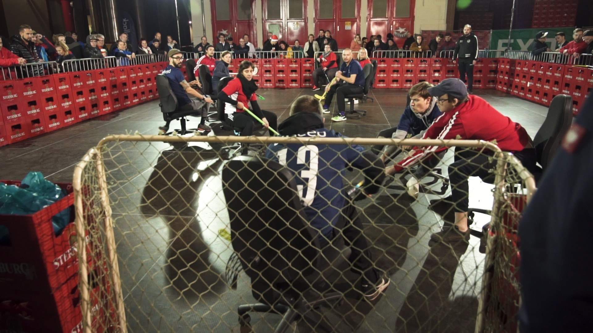 Spieler in Rollstühlen spielen Hockey in einer Halle mit Zuschauern im Hintergrund.