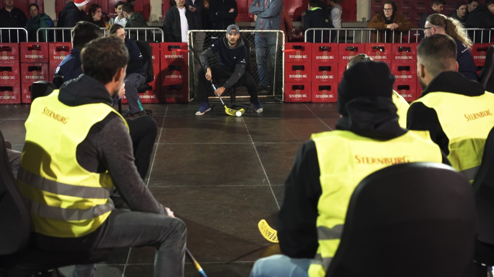 Mann sitzt auf einem Hocker und spielt Hallenhockey, Zuschauer im Hintergrund.