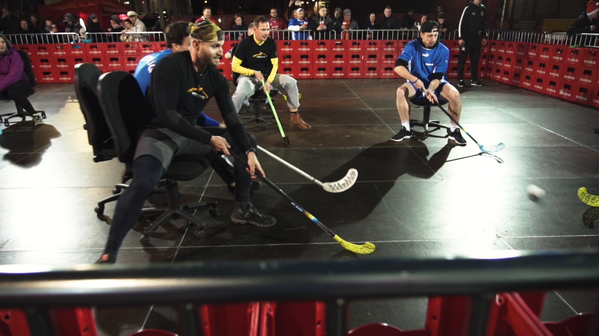 Vier Männer spielen Eishockey auf Bürostühlen in einer Halle vor Zuschauern.
