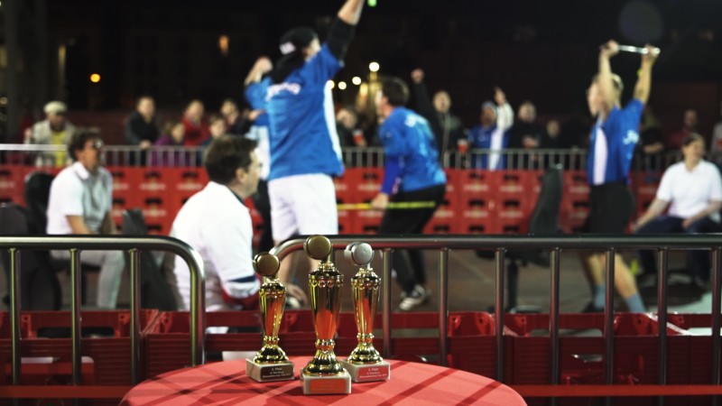 Drei Trophäen stehen im Vordergrund auf einem Tisch bei einem Sportevent. Im Hintergrund jubeln Sportler und Zuschauer.