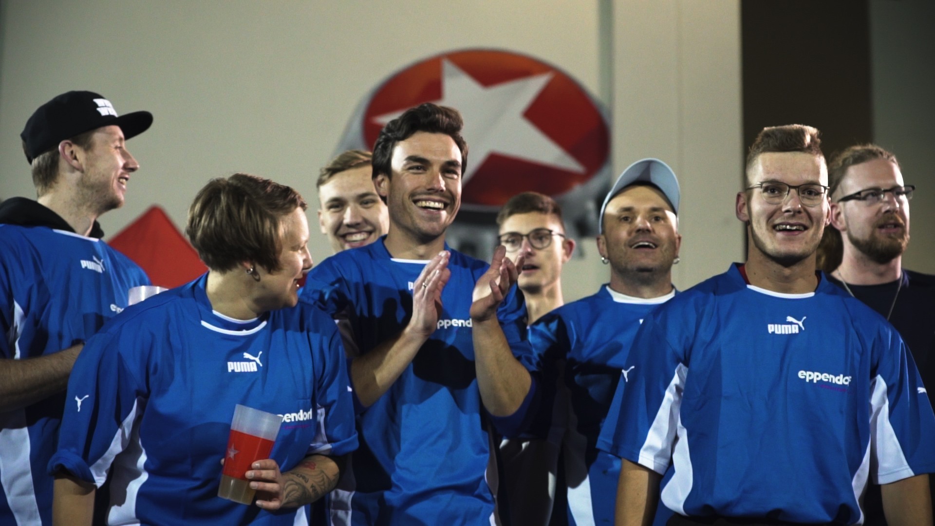 Gruppe von acht lächelnden Männern in blauen Sporttrikots klatscht Beifall.