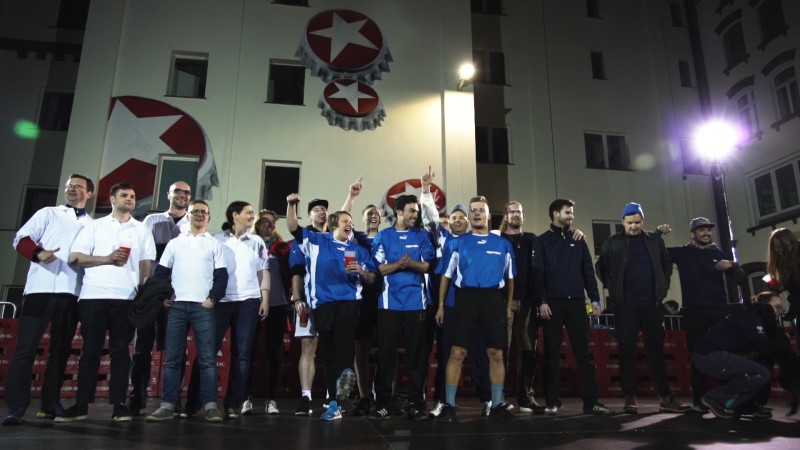 Eine Gruppe von Menschen steht lächelnd vor einem beleuchteten Gebäude bei Nacht. Einige tragen blaue oder weiße Arbeitskleidung.