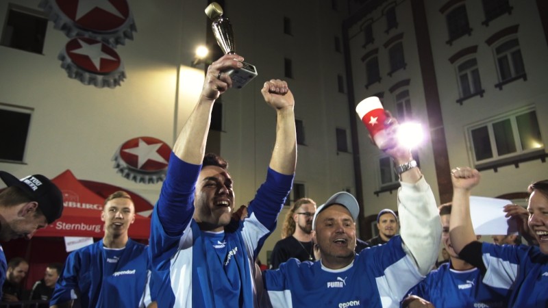 Ein glücklicher Mann in einem blauen Sporttrikot hebt einen Pokal, umgeben von jubelnden Menschen.