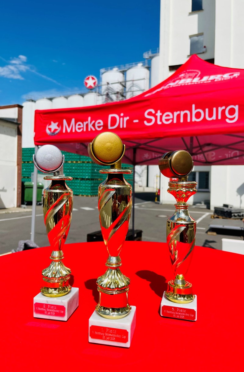 Drei Pokale stehen auf einem roten Tisch vor einem Zelt und Industriekulisse.