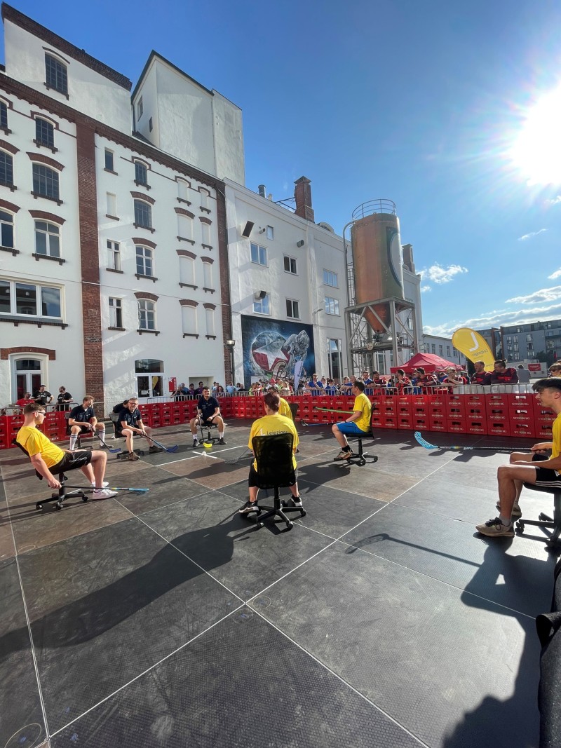 Mehrere Personen sitzen auf Bürostühlen in einer Outdoor-Veranstaltung mit Stadthintergrund.